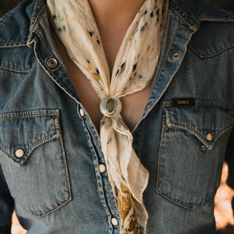 A close-up of a person wearing a light beige scarf with a subtle floral pattern and a silver-toned decorative button, tied around an open blue denim shirt.