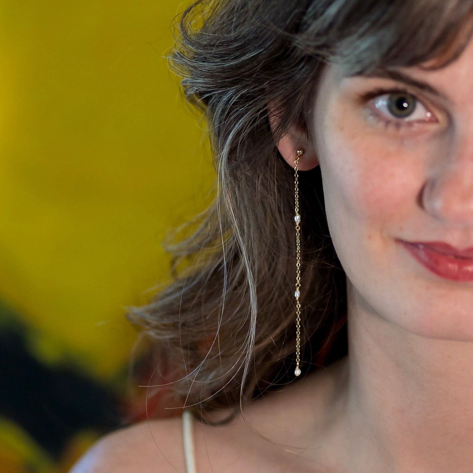 A close-up lifestyle shot of a woman wearing a delicate gold dangle earring with small white bead accents, set against a vibrant yellow background.